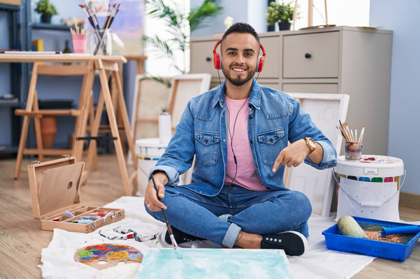 Fiatal spanyol férfi festő ül a padlón a művészeti stúdióban mosolyogva boldog rámutatva a kezét és ujját  - Fotó, kép