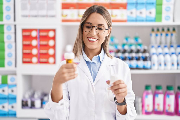 Junge schöne hispanische Apothekerin lächelt zuversichtlich und hält Tablettenflaschen in der Apotheke - Foto, Bild