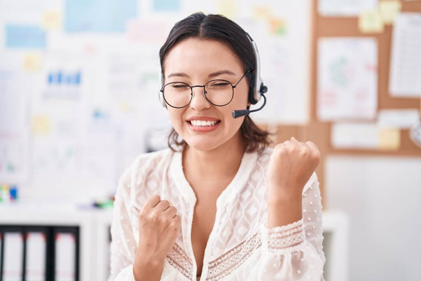 Hispanic young woman working at the office wearing headset and glasses celebrating surprised and amazed for success with arms raised and eyes closed  - Photo, image