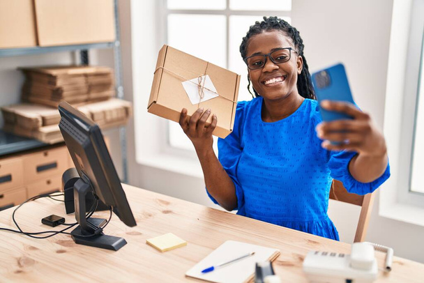 African american woman ecommerce business worker make selfie by smartphone holding package at office - Zdjęcie, obraz
