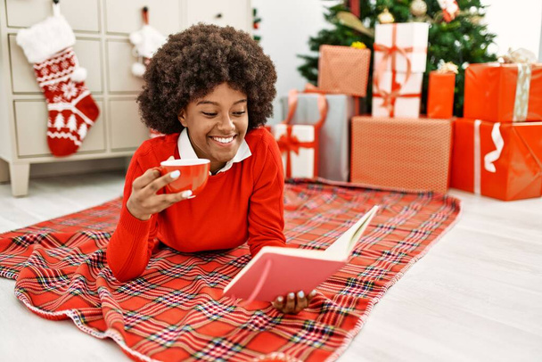 Genç Afrikalı Amerikalı kadın kahve içiyor ve kitap okuyor. Evdeki Noel ağacının yanında yatıyor. - Fotoğraf, Görsel