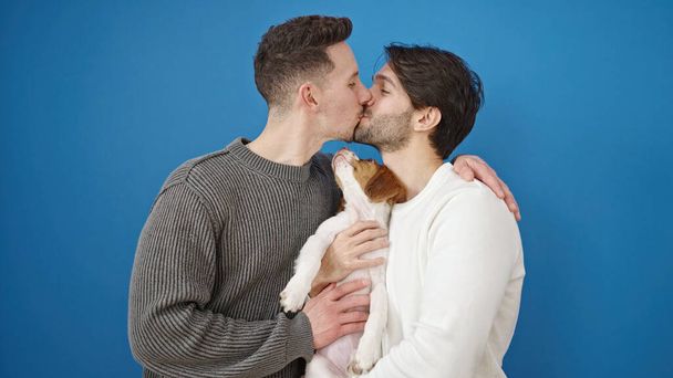 Zwei Männer Paar küsst stehend umarmenden Hund vor isoliertem blauen Hintergrund - Foto, Bild