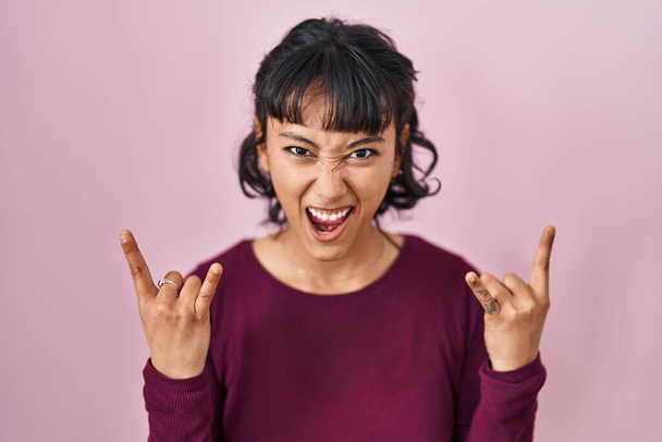 Young beautiful woman standing over pink background shouting with crazy expression doing rock symbol with hands up. music star. heavy concept.  - Photo, Image