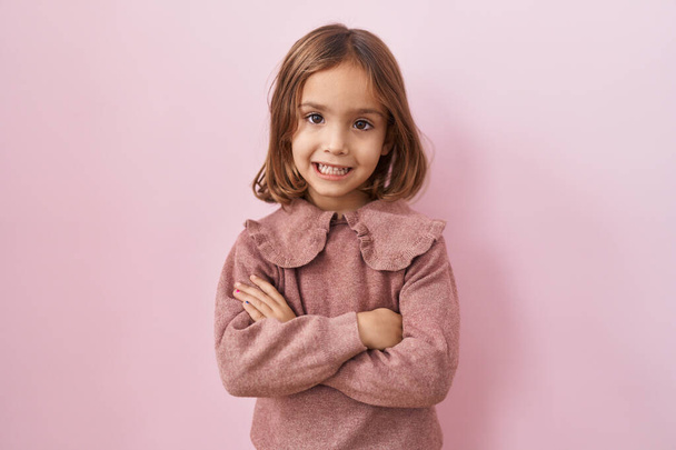 Μικρό ισπανόφωνο κορίτσι στέκεται πάνω από ροζ φόντο χαρούμενο πρόσωπο χαμογελώντας με σταυρωμένα χέρια κοιτάζοντας την κάμερα. θετικό πρόσωπο.  - Φωτογραφία, εικόνα