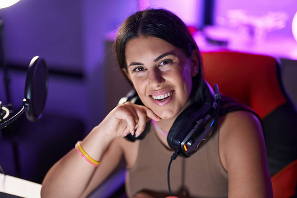 Νέα όμορφη ισπανόφωνη γυναίκα streamer χαμογελά αυτοπεποίθηση κάθεται στο τραπέζι στο gaming room - Φωτογραφία, εικόνα