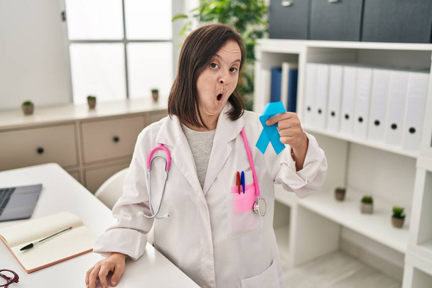 Hispanic girl with down syndrome wearing doctor uniform holding blue ribbon scared and amazed with open mouth for surprise, disbelief face  - Photo, Image