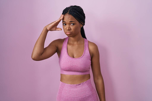 Afro-Amerikaanse vrouw met vlechten dragen sportkleding over roze achtergrond bezorgd en gestrest over een probleem met de hand op het voorhoofd, nerveus en angstig voor crisis  - Foto, afbeelding