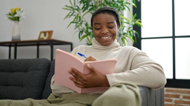 Африканська американка пише на записнику сидячи вдома на дивані. - Фото, зображення