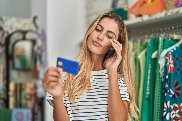 Jeune femme blonde client parlant sur smartphone tenant carte de crédit au magasin de vêtements - Photo, image