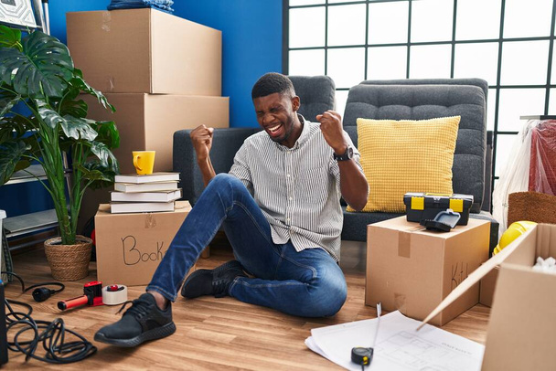 Afrikanisch-amerikanischer Mann sitzt auf dem Boden in seinem neuen Zuhause sehr glücklich und aufgeregt und macht Siegergeste mit erhobenen Armen, lächelt und schreit nach Erfolg. Festkonzept.  - Foto, Bild