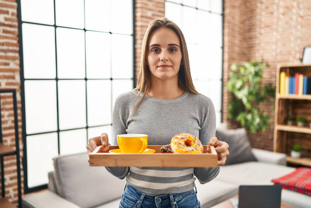 Young woman holding tray with breakfast food relaxed with serious expression on face. simple and natural looking at the camera.  - Фото, зображення