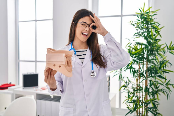 Jeune femme brune médecin tenant col cervical souriant heureux faisant signe ok avec la main sur les yeux regardant à travers les doigts  - Photo, image