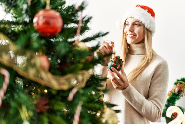 Junge blonde Frau lächelt selbstbewusst und schmückt Weihnachtsbaum zu Hause - Foto, Bild