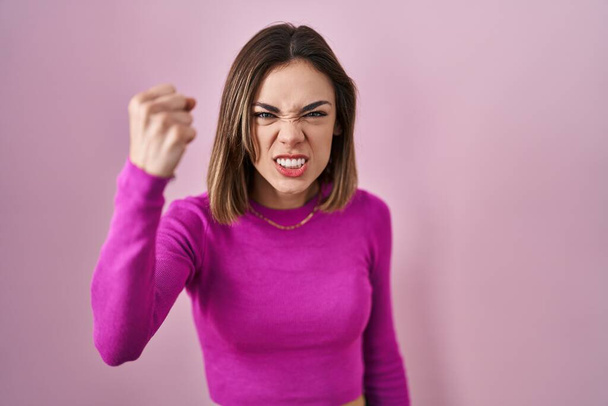 Pembe arka planda öfkeli ve kızgın duran İspanyol kadın öfkeyle bağırırken yumruğunu kaldırıyor ve öfkeleniyor. öfke ve agresif konsept.  - Fotoğraf, Görsel