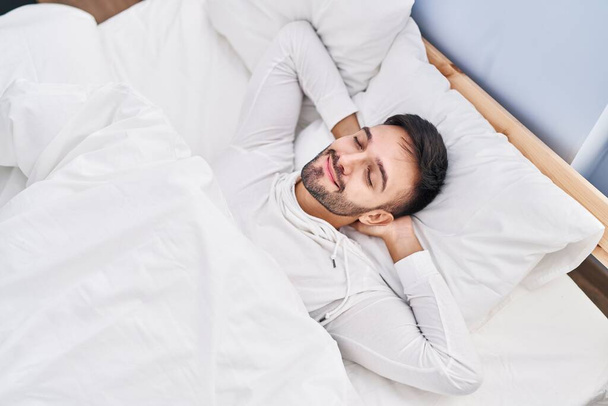 Junger hispanischer Mann lächelt selbstbewusst auf Bett im Schlafzimmer liegend - Foto, Bild