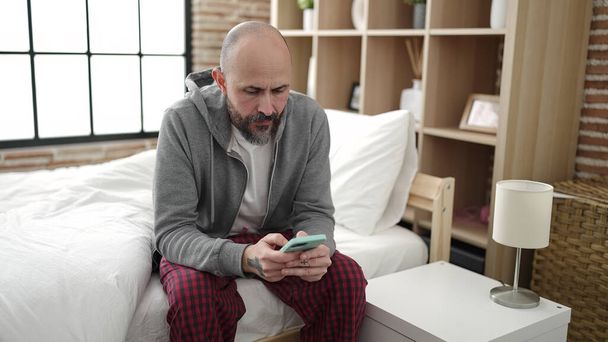 Joven hombre calvo usando teléfono inteligente sentado en la cama en el dormitorio - Foto, Imagen