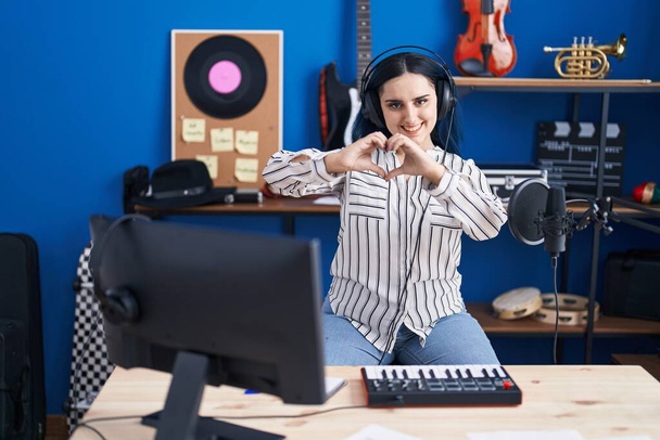 Rapariga moderna com cabelo azul no estúdio de música usando fones de ouvido sorrindo no amor fazendo forma de símbolo do coração com as mãos. conceito romântico.  - Foto, Imagem