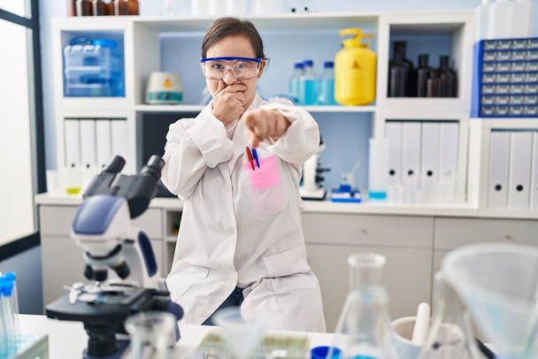 Латиноамериканка с синдромом Дауна, работающая в научной лаборатории, смеющаяся над тобой, указывающая пальцем на камеру с рукой над ртом, выражение стыда  - Фото, изображение