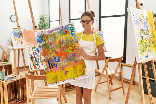 Adorable chica sonriendo confiado sosteniendo dibujo de lona en el estudio de arte - Foto, imagen