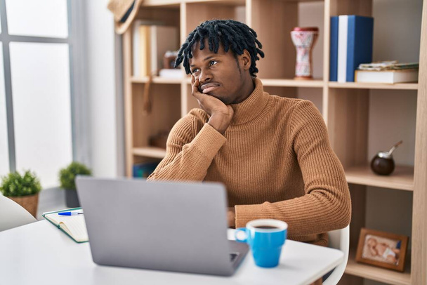 Młody Afrykanin z dredami pracujący z komputerowym laptopem wyglądający na zmęczonego i znudzonego problemami z depresją z skrzyżowanymi ramionami.  - Zdjęcie, obraz