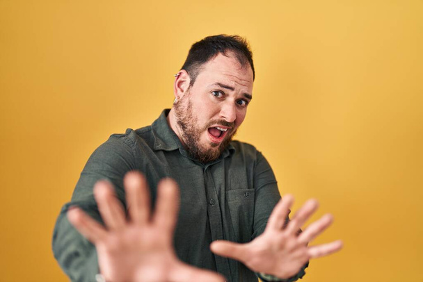 Plus taille homme hispanique avec barbe debout sur fond jaune peur et terrifié par l'expression de la peur arrêter geste avec les mains, criant en état de choc. concept de panique.  - Photo, image