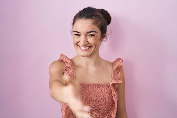 Mladá puberťačka stojí nad růžovým pozadím a usmívá se přátelsky a nabízí potřesení rukou jako pozdrav a přivítání. úspěšný obchod.  - Fotografie, Obrázek