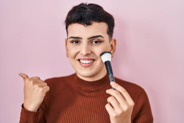 Non binary person applying make up pointing thumb up to the side smiling happy with open mouth  - Φωτογραφία, εικόνα