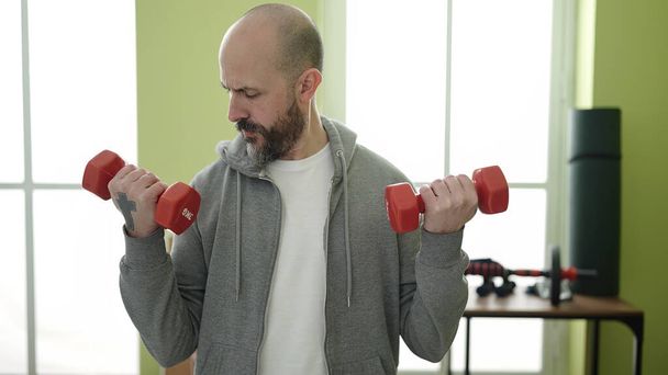 Young bald man smiling confident using dumbbells training at sport center - Photo, Image