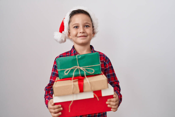 Pequeño niño hispano con sombrero de navidad sosteniendo regalos celebrando loco y sorprendido por el éxito con los ojos abiertos gritando emocionado.  - Foto, imagen