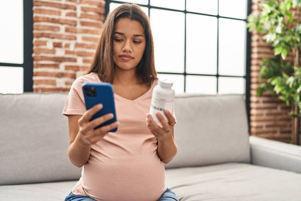 Young latin woman pregnant using smartphone holding bottle of pills at home - Fotoğraf, Görsel