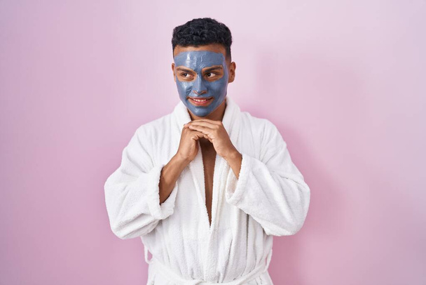 Jonge Latijns-Amerikaanse man draagt schoonheid gezichtsmasker en badjas lachen nerveus en opgewonden met handen op kin kijken naar de zijkant  - Foto, afbeelding