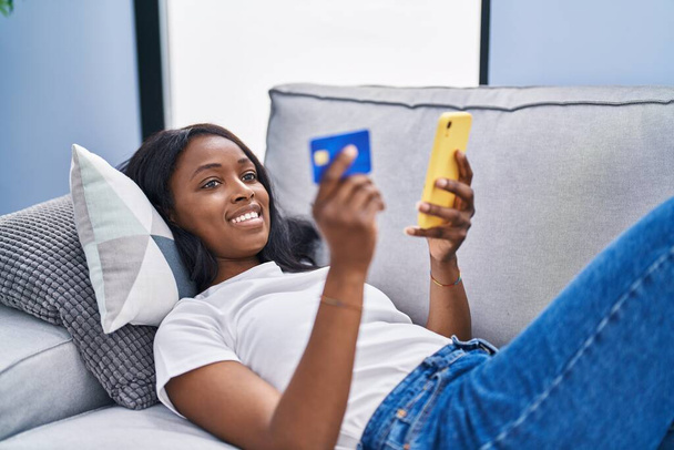 Jonge Afrikaans-Amerikaanse vrouw met behulp van smartphone en creditcard zittend op de bank thuis - Foto, afbeelding