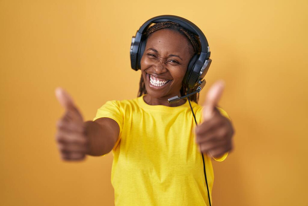 Africká americká žena, která poslouchá hudbu pomocí sluchátek, která schvaluje pozitivní gesto rukou, palce nahoru, usmívá se a je šťastná za úspěch. gesto vítěze.  - Fotografie, Obrázek