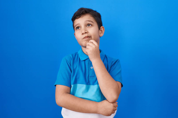 Kleine Latijns-Amerikaanse jongen die over een blauwe achtergrond staat te glimlachen en zelfverzekerd kijkt naar de camera met gekruiste armen en hand op kin. positief denken.  - Foto, afbeelding