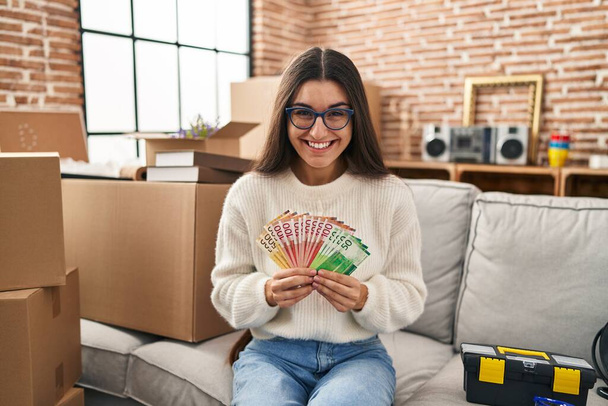Jeune femme hispanique assise sur le canapé à la nouvelle maison tenant de l'argent souriant et riant fort à haute voix parce que drôle blague folle.  - Photo, image