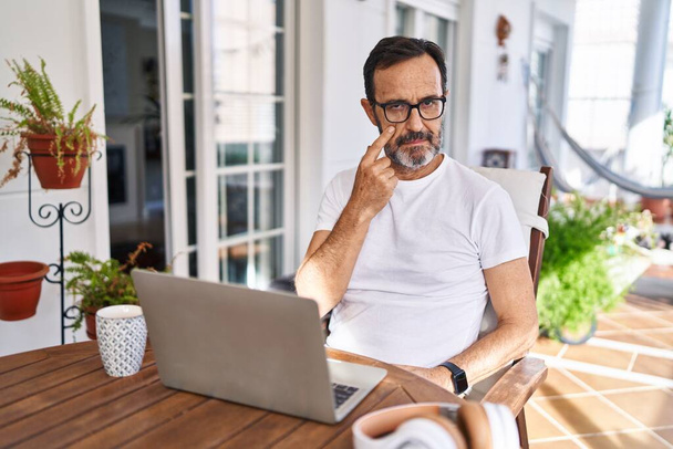 Hombre de mediana edad usando computadora portátil en casa apuntando al ojo observándote gesto, expresión sospechosa  - Foto, Imagen