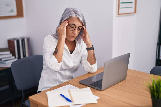 Femme d'âge moyen aux cheveux gris travailleur d'affaires stressé à l'aide d'un ordinateur portable au bureau - Photo, image