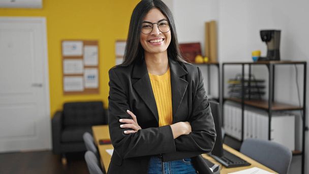 Junge schöne hispanische Geschäftsfrau lächelt selbstbewusst mit verschränkten Armen im Büro - Foto, Bild