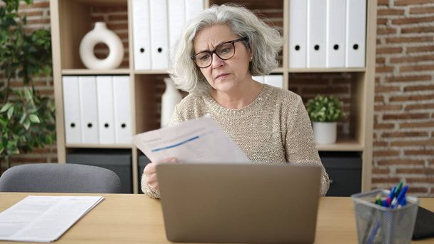 Femme d'âge moyen avec travailleur d'affaires cheveux gris en utilisant un document de lecture d'ordinateur portable au bureau - Photo, image