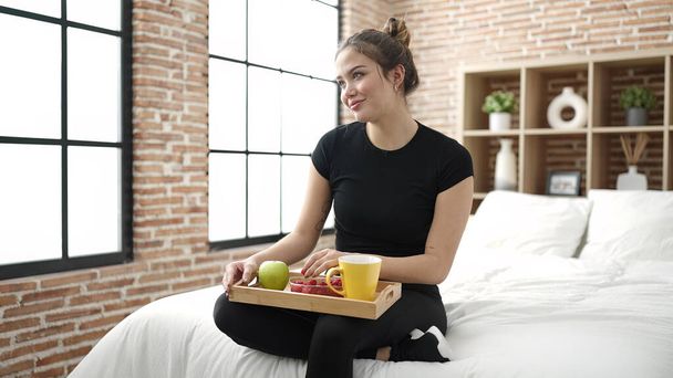 Young beautiful hispanic woman having healthy breakfast sitting on bed at bedroom - Photo, Image