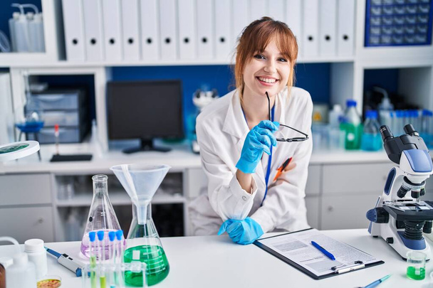 Young woman scientist smiling confident holding glasses at laboratory - Foto, Imagem