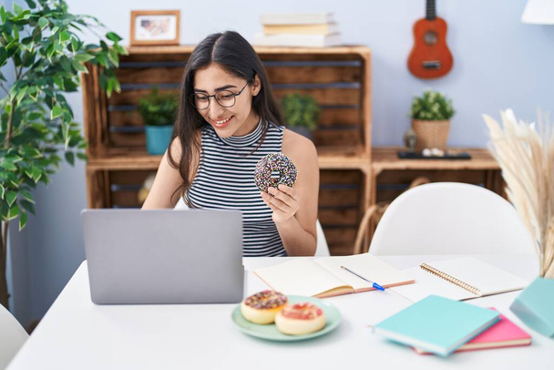 Young hispanic girl eating doughnut studying at home - Foto, afbeelding
