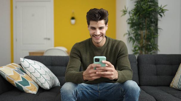 Hombre hispano joven usando teléfono inteligente sentado en el sofá en casa - Foto, Imagen