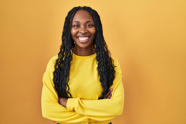 Afrikaanse vrouw staat over gele achtergrond gelukkig gezicht glimlachend met gekruiste armen kijkend naar de camera. positieve persoon.  - Foto, afbeelding