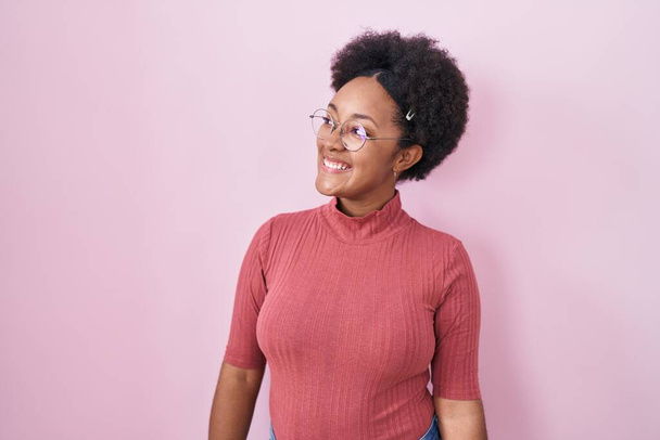Όμορφη Αφρικανή γυναίκα με σγουρά μαλλιά στέκεται πάνω από ροζ φόντο κοιτάζοντας μακριά στην πλευρά με χαμόγελο στο πρόσωπο, φυσική έκφραση. γελώντας σίγουρος.  - Φωτογραφία, εικόνα