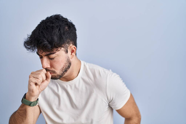Hombre hispano con barba de pie sobre fondo blanco sintiéndose mal y tosiendo como síntoma de resfriado o bronquitis. concepto de atención sanitaria.  - Foto, imagen