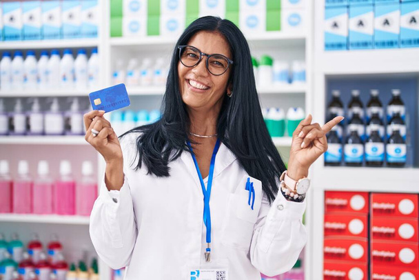 Mujer hispana madura trabajando en farmacia sosteniendo tarjeta de crédito sonriendo feliz señalando con la mano y el dedo a un lado  - Foto, Imagen