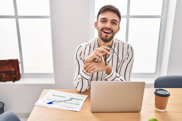 Young hispanic man business worker smiling confident sitting on table at office - Zdjęcie, obraz