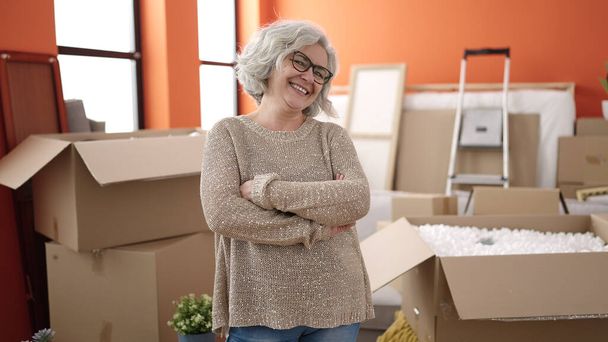 Kobieta w średnim wieku z siwymi włosami uśmiechnięta pewnie stojąca z rękoma skrzyżowanymi gestem w nowym domu - Zdjęcie, obraz