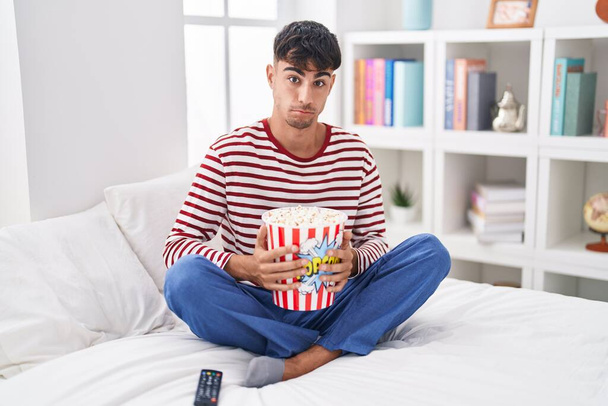 Joven hispano comiendo palomitas de maíz sentado en la cama viendo una película escéptico y nervioso, frunciendo el ceño molesto debido a un problema. persona negativa.  - Foto, imagen